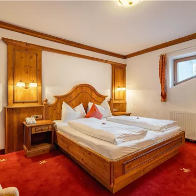 A cozy bedroom with a large double bed and wooden paneling. The room has red carpets and windows with decorative curtains.