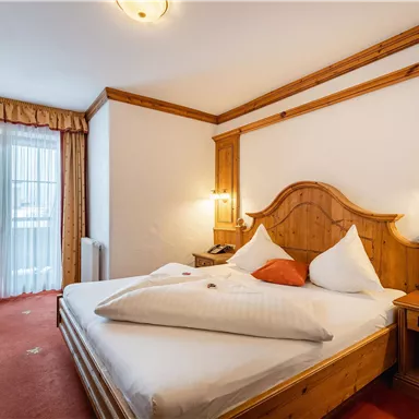 A cozy hotel room with wooden decor and a large bed. There is a table, a chair, and a window with curtains.