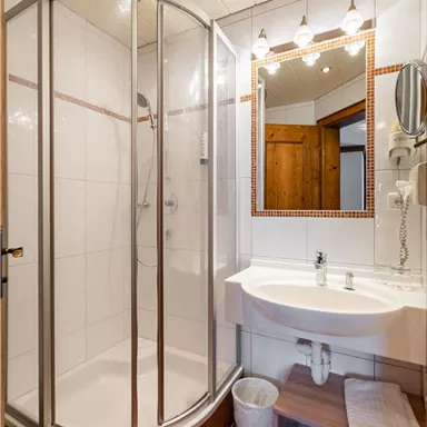 Ein modernes Badezimmer mit einer Duschkabine, einem Waschbecken und einem Spiegel. Die Wände sind hell gefliest und es gibt einen Holzschrank im Hintergrund.