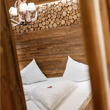 Ein gemütliches Schlafzimmer mit einem Holzbett und weißen Kissen. Die Wände sind mit Holzscheiten dekoriert, und eine elegante Lampe hängt von der Decke.
