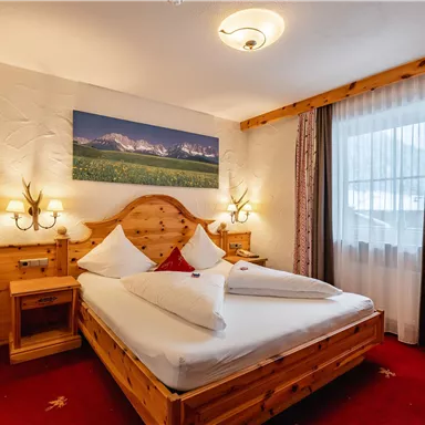 A cozy bedroom with a wooden bed and red carpet. The room is brightly lit and has large windows looking outside.