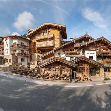 A picturesque alpine hotel with wooden balconies and an inviting terrace. In the background, green mountains and a blue sky can be seen.