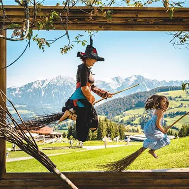 A girl and a witch are happily flying on brooms in a picturesque landscape frame. In the background, green hills and mountains are visible.