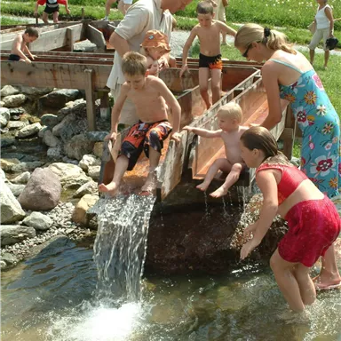 Eine Gruppe von Kindern und Erwachsenen spielt am Wasser. Sie sitzen auf einer Holzrinne und genießen den sprudelnden Wasserfluss.