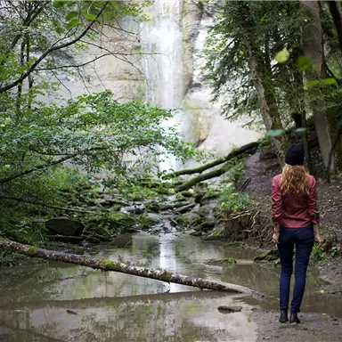 Eine Frau steht an einem kleinen Wasserfall in einem bewaldeten Gebiet. Der Wald ist grün und üppig, mit einem klaren Wasserbecken im Vordergrund.