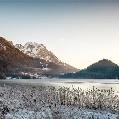 A winter landscape with a frozen lake and snow-covered mountains in the background. The sky is clear and the sun is shining gently.