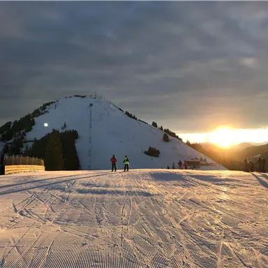 A snow-covered slope with two skiers heading into the evening sun. In the background, a wooded mountain slope and the peak of the Hohe Salve in the SkiWelt Wilder Kaiser - Brixental can be seen.