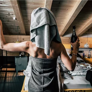 Eine Person mit einem Handtuch auf dem Kopf hält ein Glas in einer Hand und eine Flasche in der anderen. Der Raum hat eine gemütliche Atmosphäre mit Holzdetails und einer modernen Einrichtung.