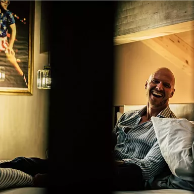 Ein lächelnder Mann sitzt auf einem Bett in einem gemütlichen Raum. Im Hintergrund hängt ein lebhaftes Bild an der Wand.