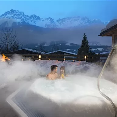 Ein romantischer Außen-Whirlpool umgeben von Nebel und Bergen im Hintergrund. Die Szene ist abends mit sanftem Licht beleuchtet.