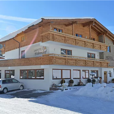 A charming hotel in winter with wooden cladding and a snow-covered ground. The surroundings are adorned with trees and the sky is clear and blue.