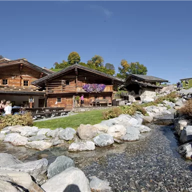 Eine malerische Almhütte mit Holzfassaden und bunten Blumen. Im Vordergrund fließt ein klarer Bach über glatte Steine.