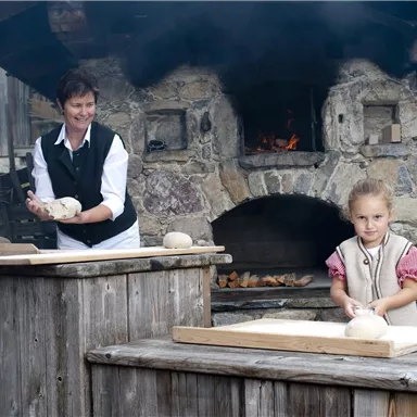 Eine Frau und ein Kind stehen an Holztischen und bereiten Teig vor. Im Hintergrund ist ein traditioneller Steinofen zu sehen.