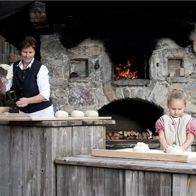 Eine Frau und ein Mädchen bereiten Teig in einer traditionellen Holzofen-Umgebung vor. Im Hintergrund ist ein offenes Feuer zu sehen.