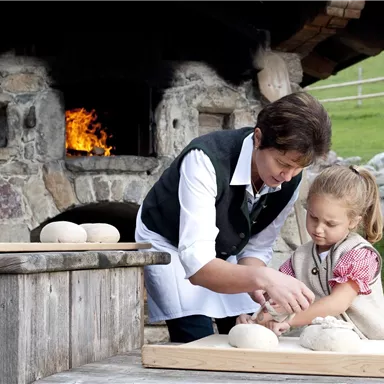 Eine Frau und ein kleines Mädchen backen gemeinsam Brot vor einem offenen Kamin. Beide arbeiten an einem Holztisch mit Teig.