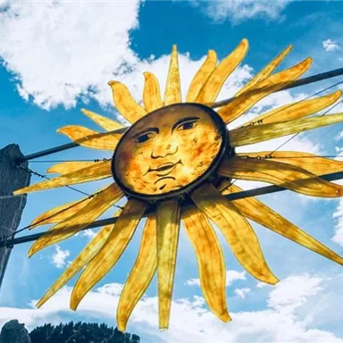 Eine sonnengelbe Sonnenskulptur vor einem blauen Himmel mit Wolken. Die Sonne hat ein freundliches Gesicht und strahlt Wärme aus.
