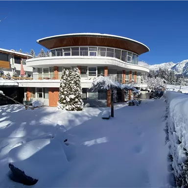 Ein modernes Haus im Schnee, umgeben von einer verschneiten Landschaft. Im Hintergrund sind Berge sichtbar und der Himmel ist klar und blau.