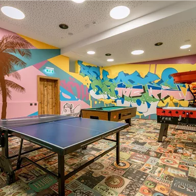 Ein bunt gestalteter Spielraum mit einem Tischtennistisch und einem Tischfußballspiel. Die Wände sind kreativ mit Palmen und farbenfrohen Grafiken dekoriert.