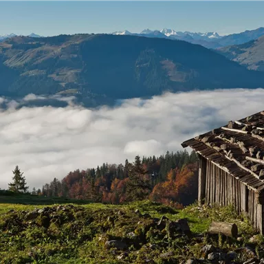 Eine Aussicht auf hohe Berge, umgeben von Wolken. Im Vordergrund steht ein altes Holzgebäude auf einer grünen Wiese.