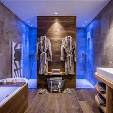 Ein modernes Badezimmer mit einer Badewanne und einer eleganten Dusche. Zwei Bademäntel hängen an der Wand, umgeben von stilvollen Holz- und Fliesenelementen.
