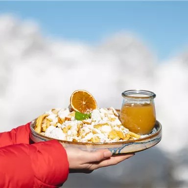 A plate of freshly prepared Kaiserschmarrn with an orange slice, accompanied by a glass of fruit juice. In the background, snow-covered mountains can be seen.
