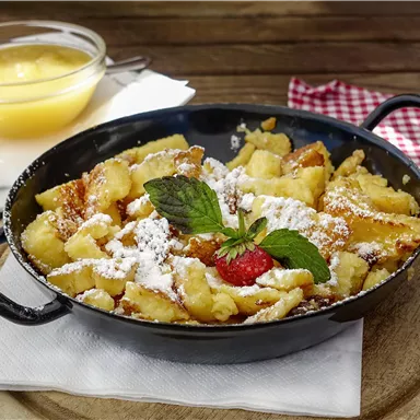A delicious Kaiserschmarrn in a black pan, garnished with powdered sugar and a fresh mint leaf. Next to it is a bowl of applesauce.
