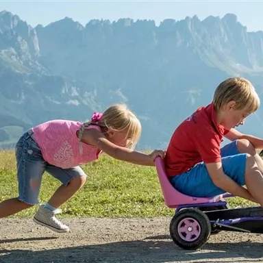 Ein Junge fährt auf einem kleinen Fahrzeug, während ein Mädchen ihn von hinten schiebt. Die Szene spielt vor einer beeindruckenden Bergkulisse.