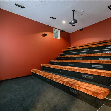 Ein modernes Heimkino mit mehreren Ebenen von bequemen Sitzen. Die Wände sind in einem warmen Rotton gestrichen, und es gibt eine Leinwand für Filmvorführungen.