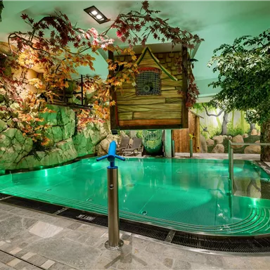 A stylish indoor pool with green water and natural decoration. Surrounded by artificial trees and rocks, the environment creates a relaxing atmosphere.