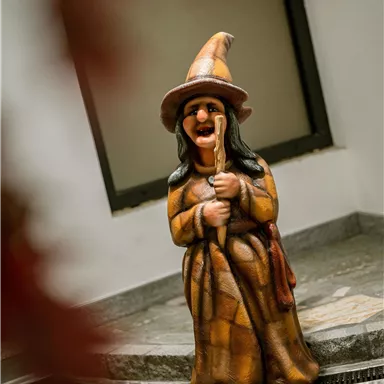 Eine fröhliche Hexenfigur in einem braunen Kleid steht neben einem Wasserspiel. Sie hält einen Besen und lächelt.