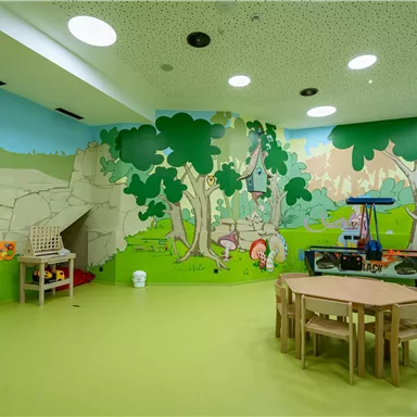Ein bunter Spielraum mit einer fantasievollen Wandmalerei von Tieren und Natur. Es gibt mehrere Holztische und Stühle für Kinder.
