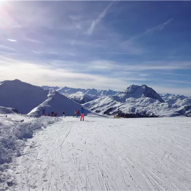 Eine schneebedeckte Berglandschaft unter einem klaren blauen Himmel. Skifahrer genießen die Pisten in der Ferne.