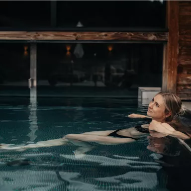Eine Frau entspannt im Wasser eines Innenpools. Die Umgebung ist modern und einladend, mit warmem Licht und Holzdetails.