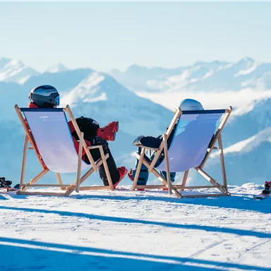 Zwei Personen sitzen entspannt auf Liegestühlen in den Bergen. Im Hintergrund sind schneebedeckte Gipfel und ein klarer Himmel zu sehen.