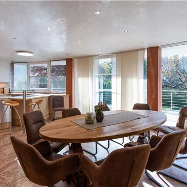 Moderne Wohnküche mit einem großen Esstisch und gemütlichen Stühlen. Viel Tageslicht durch große Fenster sorgt für eine einladende Atmosphäre.