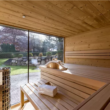 Ein modernes Saunainneres aus Holz mit Bänken und einem großen Fenster. Draussen sind Bäume und ein gepflegter Garten sichtbar.