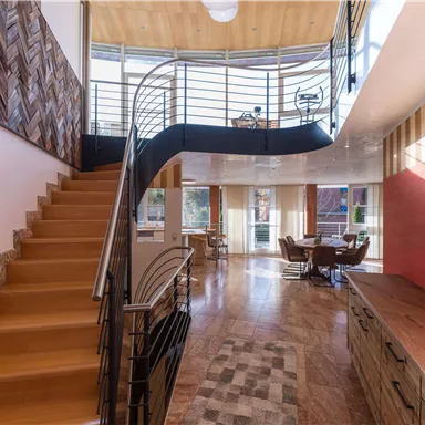 Ein modernes und stilvolles Wohnzimmer mit einer offenen Treppe aus Holz. Große Fenster lassen viel Licht herein und schaffen eine einladende Atmosphäre.