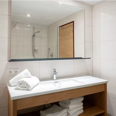 Ein modernes Badezimmer mit einem Waschbecken, einem großen Spiegel und frischen Handtüchern. Die Wände sind hell gefliest und es gibt eine Dusche im Hintergrund.