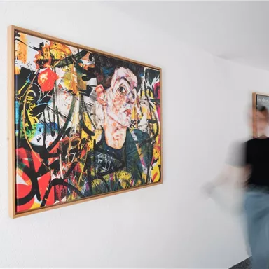 Ein lebhaftes Kunstwerk an einer Wand mit bunten Farben und dynamischen Mustern. Eine Person geht an dem Bild vorbei und schafft eine Verbindung zwischen Kunst und Bewegung.