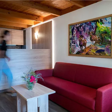 Ein gemütliches Wohnzimmer mit einem roten Sofa und einem Holztisch. An der Wand hängt ein farbenfrohes Gemälde, während eine Person vorbeigeht.
