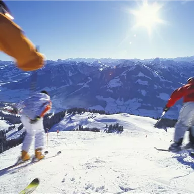 Ein malerischer Blick auf eine schneebedeckte Berglandschaft. Zwei Skifahrer fahren die Piste hinunter, während die Sonne am klaren Himmel scheint.