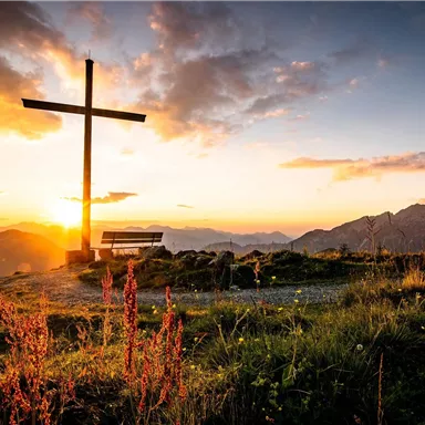 A large cross stands on a hill at twilight. The sun sets behind the mountains, painting the sky in warm hues.