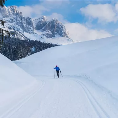 Ein Skifahrer läuft durch eine schneebedeckte Landschaft. Im Hintergrund sind majestätische Berge und ein klarer blauer Himmel zu sehen.