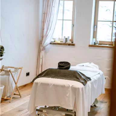 A quiet wellness room with a treatment table and large windows. The wooden floor and soft colors create a relaxed atmosphere.