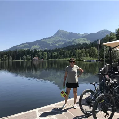 Ein ruhiger See umgeben von Bergen und dichtem Wald. Im Vordergrund steht eine Person mit einem gelben Schwimmspielzeug und Fahrrädern.