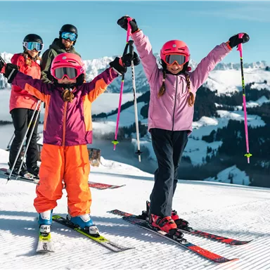 A group of skiers is standing on a snow-covered slope. Two excited children in colorful ski outfits are raising their hands in the air.