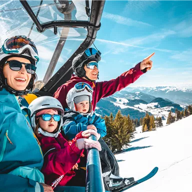 A family in a ski lift enjoying the view of the snow-covered mountains. They are wearing ski helmets and goggles and appear cheerful.