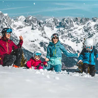 A family is playing in the snow with snow-covered mountains in the background. Everyone is wearing ski helmets and colorful winter clothing.