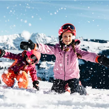 Three children are enthusiastically playing in the snow. They are laughing, throwing snowballs into the air, and wearing colorful ski clothing.