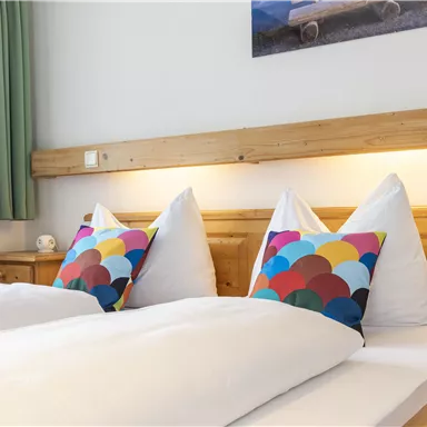 Ein gemütliches Zimmer mit zwei Einzelbetten und bunten Kissen. Die warme Holzoptik und die grünen Vorhänge schaffen eine einladende Atmosphäre.
